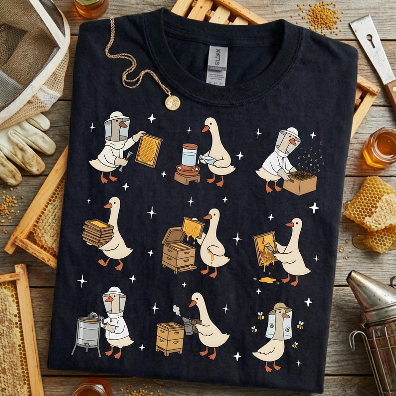 Puede incluir: Camiseta azul marino oscuro con una ilustraci&oacute;n caprichosa de gansos vestidos de apicultores. Los gansos se muestran trabajando con panales, tarros y otros equipos de apicultura. El dise&ntilde;o incluye peque&ntilde;as estrellas blancas.