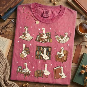 Puede incluir: Camiseta rosa descolorida con un dise&ntilde;o caprichoso de gansos de dibujos animados leyendo libros en varios escenarios. Los gansos se representan con gafas y leyendo en una hamaca, sobre pilas de libros y en un escritorio. La camiseta tiene un aspecto vintage.
