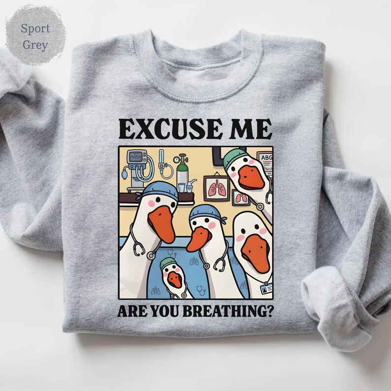 May include: Sport grey sweatshirt featuring a humorous graphic. The graphic shows cartoon geese dressed as medical professionals with stethoscopes, with the text "EXCUSE ME ARE YOU BREATHING?" in black. The geese have orange beaks and are wearing blue caps.