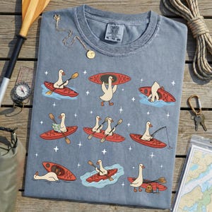 May include: A blue Comfort Colors t-shirt featuring a whimsical design of cartoon geese kayaking in red kayaks. The geese are depicted paddling, fishing, and carrying kayaks. The shirt has a crew neck and short sleeves.