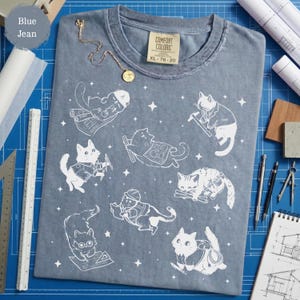 May include: Blue jean Comfort Colors t-shirt featuring white illustrations of cats in various architect roles. The shirt has a crew neck and a tag that reads XL 78-20. A gold necklace with a pendant is draped on the shirt.