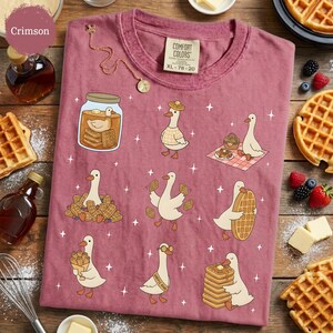 May include: Crimson Comfort Colors t-shirt with a whimsical design of cartoon geese interacting with waffles. The geese are depicted in various poses, some holding waffles, others in a jar, and some wearing hats. The shirt is size XL, 78-20.