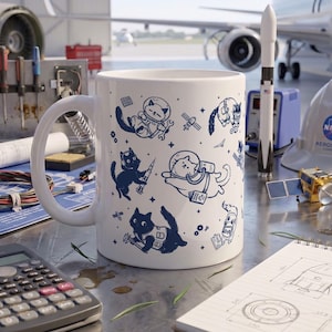 May include: A white ceramic mug featuring a repeating dark blue graphic print of various cats in space suits, floating with tools, and interacting with rockets and satellites. The design includes small stars and geometric shapes scattered across the surface.