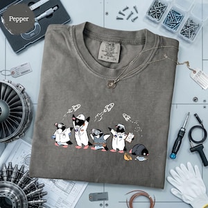 May include: A charcoal gray t-shirt featuring a graphic print of five cartoon penguins dressed as space engineers and astronauts, with small white rockets flying above them. The shirt has a crew neckline and a Comfort Colors brand label inside.