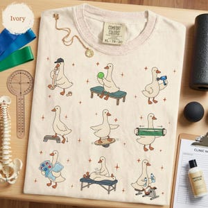 May include: An ivory t-shirt featuring a graphic print of nine white ducks performing various physical therapy exercises, including using a foam roller, balance board, and resistance bands, with small brown star accents scattered across the fabric.