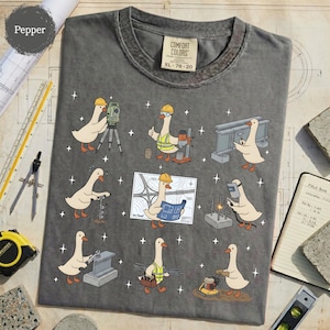 May include: Pepper-colored Comfort Colors t-shirt with a graphic of cartoon geese in construction worker attire. The geese are depicted using surveying equipment, drilling, welding, and reading blueprints. The shirt has a crew neck and a tag that reads XL 78-20.
