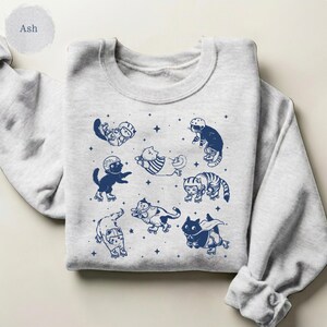 May include: A light grey sweatshirt with a crew neck. The sweatshirt features a navy blue graphic of cats wearing helmets and roller skates. The word "Ash" is printed on the upper left side of the sweatshirt.