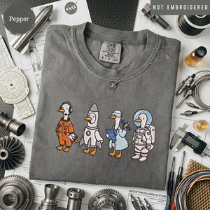 May include: A charcoal gray t-shirt featuring a graphic of four cartoon ducks dressed as space explorers, including an astronaut, a scientist, a rocket, and a pilot. The shirt has a crew neckline and a Comfort Colors brand label at the neck.