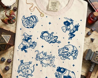 May include: An ivory t-shirt featuring a dark blue graphic print of various frogs depicted as fantasy characters, including a knight with a sword and shield, a wizard with a book, a bard with a lute, and an archer, all surrounded by small stars.