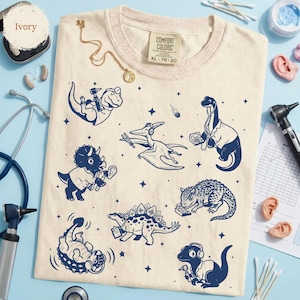 May include: An ivory-colored t-shirt featuring a dark blue graphic print of various dinosaurs dressed as medical professionals, including a T-Rex with an otoscope, a Pterodactyl with a tuning fork, and a Stegosaurus holding a medical chart.
