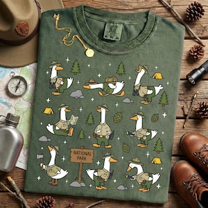 Op de afbeelding: Groen t-shirt met cartoon ganzen gekleed als parkwachters, met bomen, tenten en een "National Park" bord. De afbeelding heeft een vintage uitstraling, met een kompas, hoed en laarzen op de achtergrond.