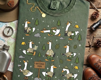 Può includere: T-shirt verde con oche dei cartoni animati vestite da ranger, con alberi, tende e un cartello "National Park". L'immagine ha un aspetto vintage, con una bussola, un cappello e stivali sullo sfondo.