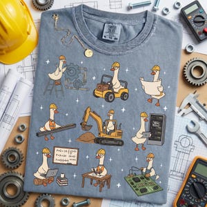 May include: A blue Comfort Colors t-shirt featuring cartoon geese in construction and engineering roles. The geese are wearing hard hats and working with machinery, blueprints, and computers. The shirt has the text "Comfort Colors" and size information.