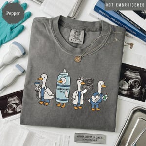 May include: A charcoal gray t-shirt featuring a graphic of four cartoon ducks dressed as medical professionals, including one as an ultrasound gel bottle. The shirt has a crew neck and a Comfort Colors label. The design is printed, not embroidered.