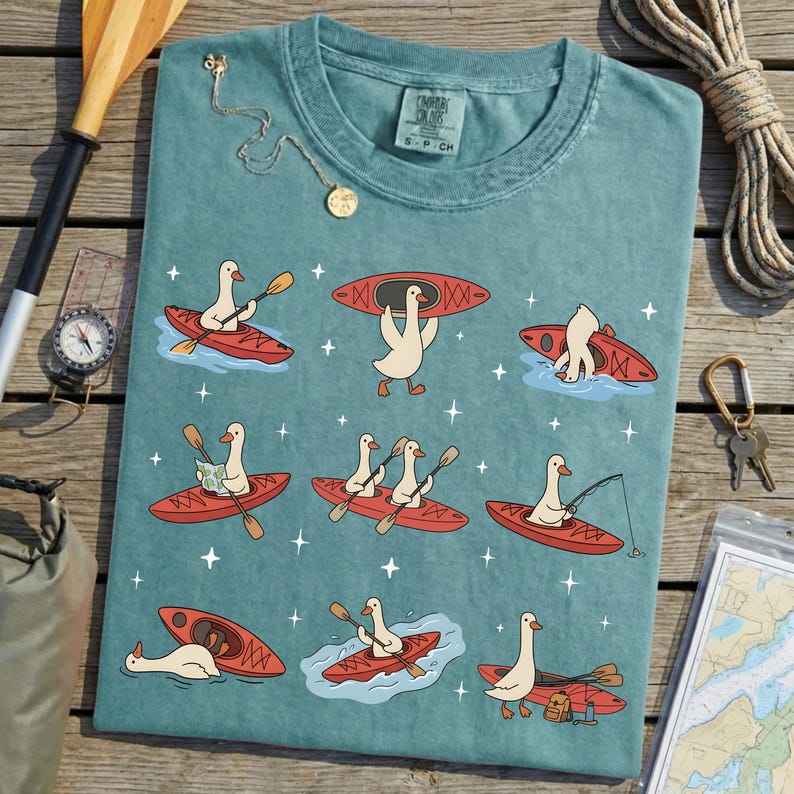 May include: Teal-colored t-shirt featuring a whimsical design of geese kayaking in red kayaks. The geese are depicted in various poses, paddling, fishing, and carrying gear. The shirt has the text "CAMPER CLUB" on the upper right.