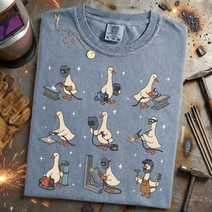 May include: A blue-grey t-shirt featuring a repeating pattern of cartoon geese welding. Each goose is depicted with welding gear, working on different metal structures. The shirt includes a Comfort Colors tag and a gold necklace.