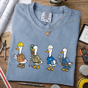 May include: A light blue Comfort Colors t-shirt featuring four cartoon geese dressed as construction workers. Each goose wears a different outfit, including a hard hat, safety vest, and overalls. The shirt has a crew neck and short sleeves.