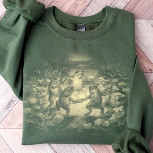 May include: A dark green sweatshirt featuring a whimsical illustration of several frogs dancing in a circle. The frogs are depicted in a vintage style, surrounded by lily pads and mushrooms. The sweatshirt has long sleeves and a crew neck.