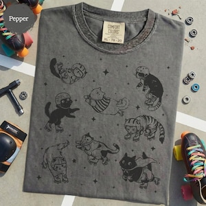 May include: A grey Comfort Colors t-shirt with a fun design of cats on roller skates. The cats are shown in different poses, wearing helmets and protective gear. The shirt has a vintage appearance, with the size XL 78-20 printed on the label.