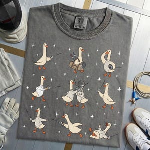 May include: Gray t-shirt featuring cartoon geese engaged in fencing, with various poses and equipment. The design includes white stars and the text "Urban Gear" on the tag. Accessories like a fencing mask, glove, and shoes are visible.