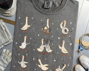 Könnte beinhalten: Graues T-Shirt mit Cartoon-Gänsen beim Fechten, in verschiedenen Posen und mit Ausrüstung. Das Design enthält weiße Sterne und den Text "Urban Gear" auf dem Etikett. Fechtmaske, Handschuh und Schuhe sind ebenfalls sichtbar.