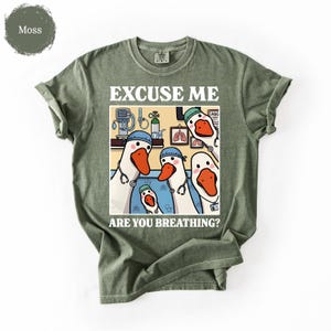 May include: Moss green t-shirt featuring a cartoon illustration of geese dressed as medical professionals. The text "EXCUSE ME ARE YOU BREATHING?" is printed above and below the illustration. The shirt has a crew neck and short sleeves.