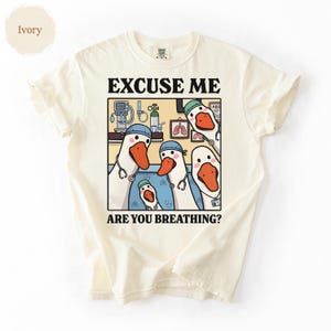 May include: Ivory-colored t-shirt featuring a cartoon illustration of geese dressed as medical professionals. The image includes the text "EXCUSE ME ARE YOU BREATHING?" The geese are wearing blue caps and stethoscopes, with medical equipment in the background.