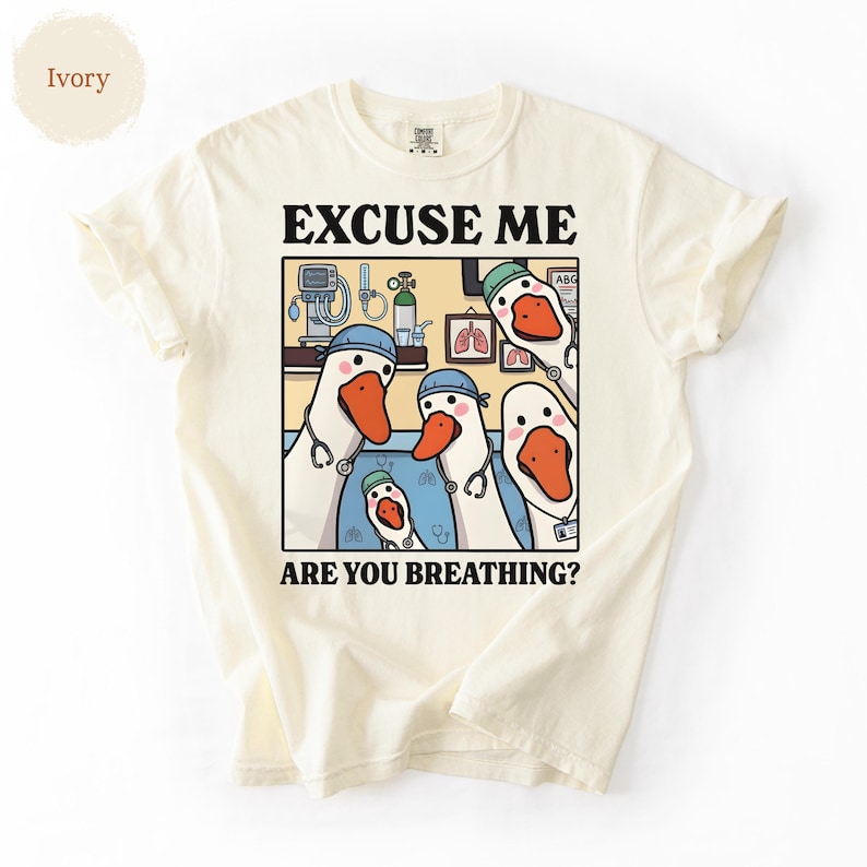 May include: Ivory-colored t-shirt featuring a cartoon illustration of geese dressed as medical professionals. The image includes the text "EXCUSE ME ARE YOU BREATHING?" The geese are wearing blue caps and stethoscopes, with medical equipment in the background.