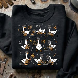 Pode incluir: Camisola preta com um padr&atilde;o repetido de gansos de desenho animado a soldar. Cada ganso &eacute; retratado com uma m&aacute;scara de soldador e um avental, segurando ferramentas e criando fa&iacute;scas. O design inclui estrelas, engrenagens e outros elementos relacionados com a soldagem.