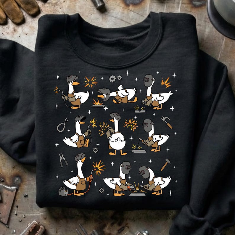 Pode incluir: Camisola preta com um padr&atilde;o repetido de gansos de desenho animado a soldar. Cada ganso &eacute; retratado com uma m&aacute;scara de soldador e um avental, segurando ferramentas e criando fa&iacute;scas. O design inclui estrelas, engrenagens e outros elementos relacionados com a soldagem.