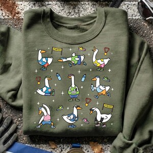 May include: Olive green sweatshirt featuring a cartoon design of geese participating in a marathon. The geese are depicted in various running poses, wearing running gear, and holding medals. The design includes water bottles, shoes, and finish line banners.