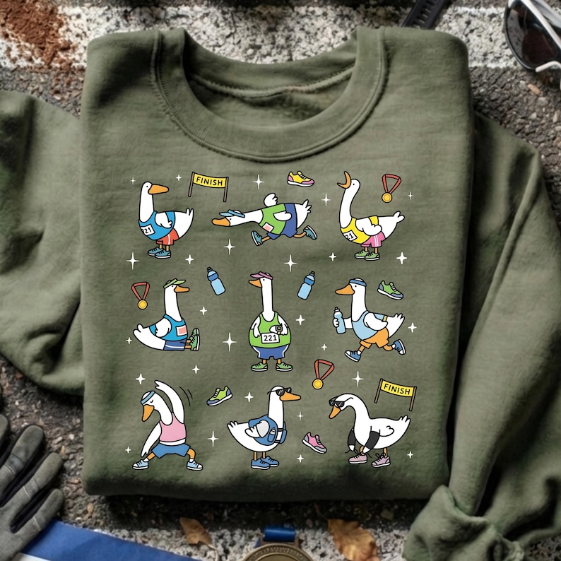May include: Olive green sweatshirt featuring a cartoon design of geese participating in a marathon. The geese are depicted in various running poses, wearing running gear, and holding medals. The design includes water bottles, shoes, and finish line banners.