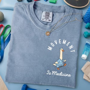 May include: A light blue Comfort Colors t-shirt with the words "MOVEMENT Is Medicine" and a cartoon goose in a doctor's uniform. A gold necklace with a pendant is on the shirt. The shirt has a small tag.