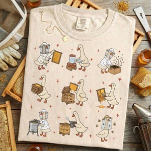 Puede incluir: Camiseta color crema con una ilustraci&oacute;n caprichosa de gansos vestidos de apicultores. El dise&ntilde;o incluye varias escenas de los gansos trabajando con panales, colmenas y miel. La camiseta tiene el texto "Champion Colors" e informaci&oacute;n de tallas.
