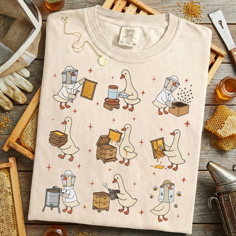 Puede incluir: Camiseta color crema con una ilustraci&oacute;n caprichosa de gansos vestidos de apicultores. El dise&ntilde;o incluye varias escenas de los gansos trabajando con panales, colmenas y miel. La camiseta tiene el texto "Champion Colors" e informaci&oacute;n de tallas.