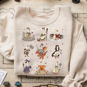 May include: Cream-colored sweatshirt featuring a whimsical design of cartoon geese in various fantasy roles. The geese are depicted as a bard, druid, scholar, warrior, knight, rogue, wizard, monk, and cleric. The design includes small red crosses.