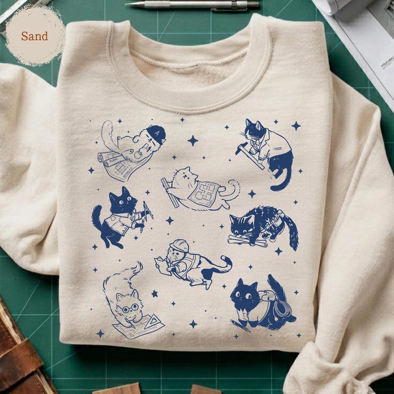 May include: Sand-colored sweatshirt featuring a repeating pattern of blue cat illustrations. Each cat is depicted in a different profession, such as an architect, construction worker, and surveyor. The design includes small blue star accents.