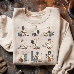 May include: Beige sweatshirt featuring a repeating pattern of cartoon geese welding. Each goose is depicted with welding gear, working on different metal structures. The design includes sparks and small red crosses, creating a fun, industrial theme.