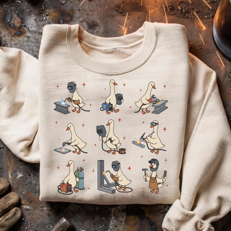 May include: Beige sweatshirt featuring a repeating pattern of cartoon geese welding. Each goose is depicted with welding gear, working on different metal structures. The design includes sparks and small red crosses, creating a fun, industrial theme.