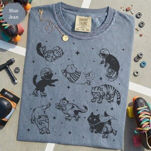 May include: Blue jean Comfort Colors t-shirt with a design of cats on roller skates and stars. The shirt has a crew neck and short sleeves. A gold necklace and roller skates are next to the shirt.