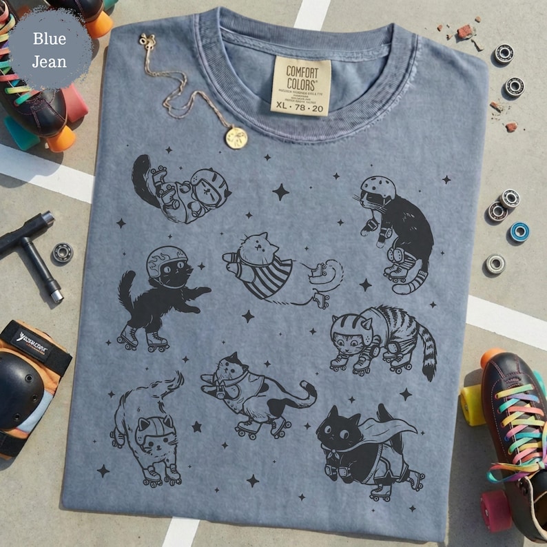 May include: Blue jean Comfort Colors t-shirt with a design of cats on roller skates and stars. The shirt has a crew neck and short sleeves. A gold necklace and roller skates are next to the shirt.