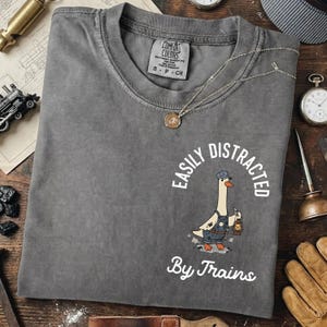 May include: Gray Comfort Colors t-shirt with the phrase "Easily Distracted By Trains" and a cartoon goose wearing a conductor's hat and overalls. The shirt has a crew neck and short sleeves. A silver necklace with a pendant is on the shirt.