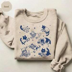 May include: A sand-colored crewneck sweatshirt featuring a dark blue graphic print of multiple cats in various poses, some wearing glasses or lab coats, surrounded by small star shapes and books, with the word Sand printed in the top corner.