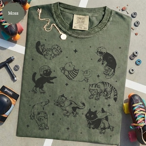May include: A moss green t-shirt featuring a design of cats on roller skates, wearing helmets. The shirt has a crew neck and a label that reads "COMFORT COLORS XL 78-20". A gold necklace and roller skates are visible.