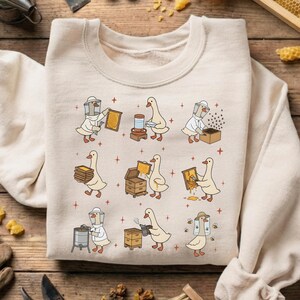 Puede incluir: Sudadera beige de cuello redondo con una ilustraci&oacute;n caprichosa de gansos apicultores. Los gansos se representan en diversas actividades ap&iacute;colas, como la recolecci&oacute;n de panales y el trabajo con miel. El dise&ntilde;o incluye acentos de estrellas rojas.