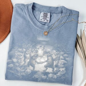 May include: A blue Comfort Colors t-shirt featuring a whimsical illustration of frogs dancing in a circle. The shirt has a small tag at the neckline and is paired with a delicate gold necklace. The shirt is folded neatly.