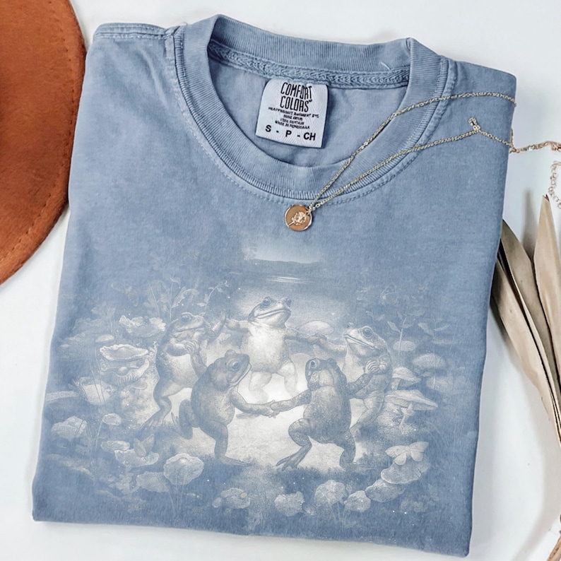 May include: A blue Comfort Colors t-shirt featuring a whimsical illustration of frogs dancing in a circle. The shirt has a small tag at the neckline and is paired with a delicate gold necklace. The shirt is folded neatly.