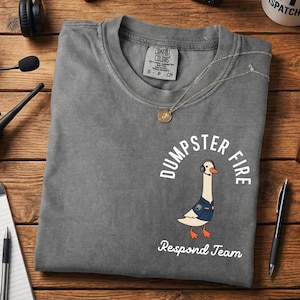 May include: A gray t-shirt with the words "Dumpster Fire" and "Respond Team" printed on it. The shirt features a cartoon goose wearing a headset and a blue vest. A silver necklace is draped on the shirt.