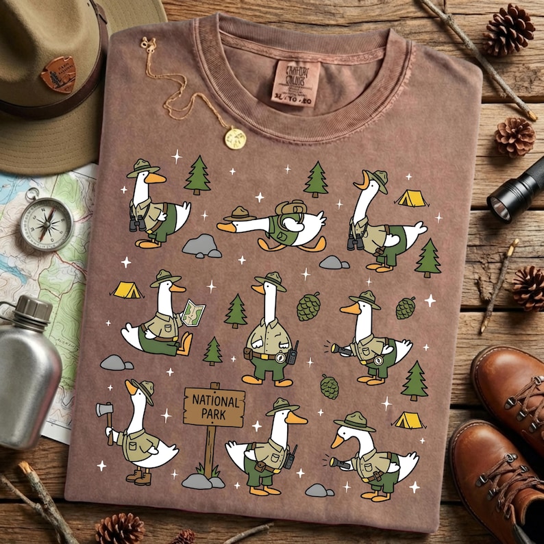 Op de afbeelding: Bruin t-shirt met een speels ontwerp van ganzen in parkwachteruniformen. De ganzen worden getoond in verschillende buitenscenario's, met bomen, tenten en een "NATIONAL PARK"-bord. Het shirt heeft een vintage uitstraling en is gemaakt van comfortabel materiaal.