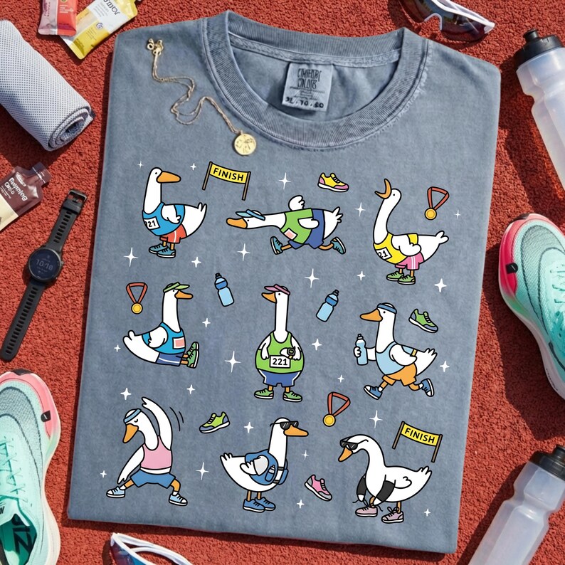 May include: A blue Comfort Colors t-shirt featuring cartoon geese in various running poses. The geese wear running gear and bibs with the number 221. The shirt includes the word "FINISH" on a banner and is surrounded by running shoes, water bottles, and medals.